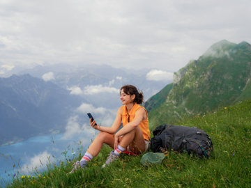 Eine junge Frau sitzt auf einer Wiese auf der Spitze eines Gipfels und blickt auf ihr Handy.